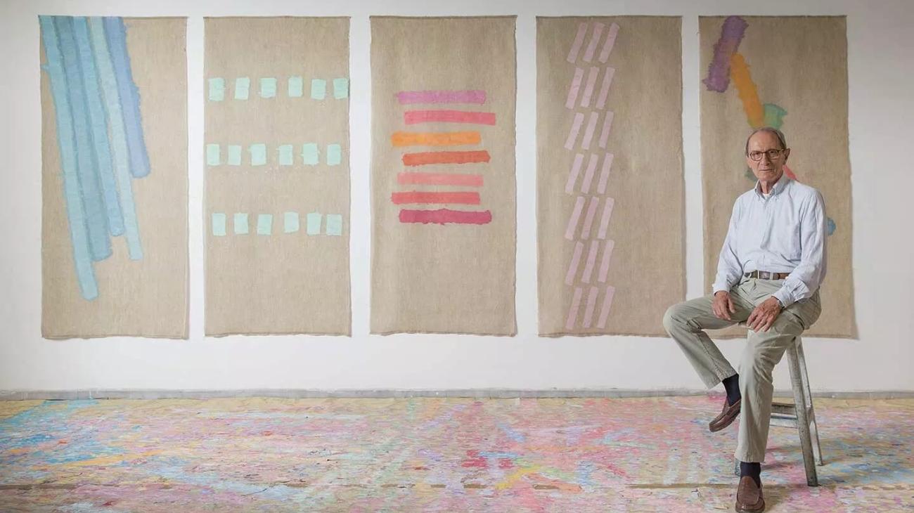 Griffa sitting on a stool in his studio with four paintings hung on the wall behind him