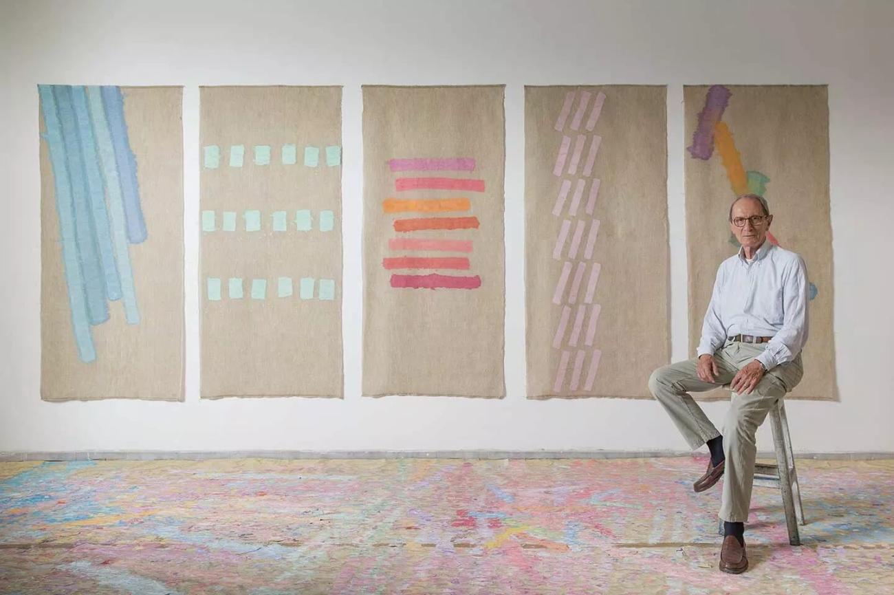 Griffa sitting on a stool in his studio with four paintings hung on the wall behind him